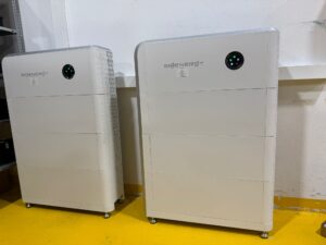 Zwei weiße Sigenergy SigenStor Energiespeichersysteme mit markantem LED-Leuchtring auf gelbem Industrieboden, bestehend aus stapelbaren Batteriemodulen und integriertem KI-Energiemanagement.